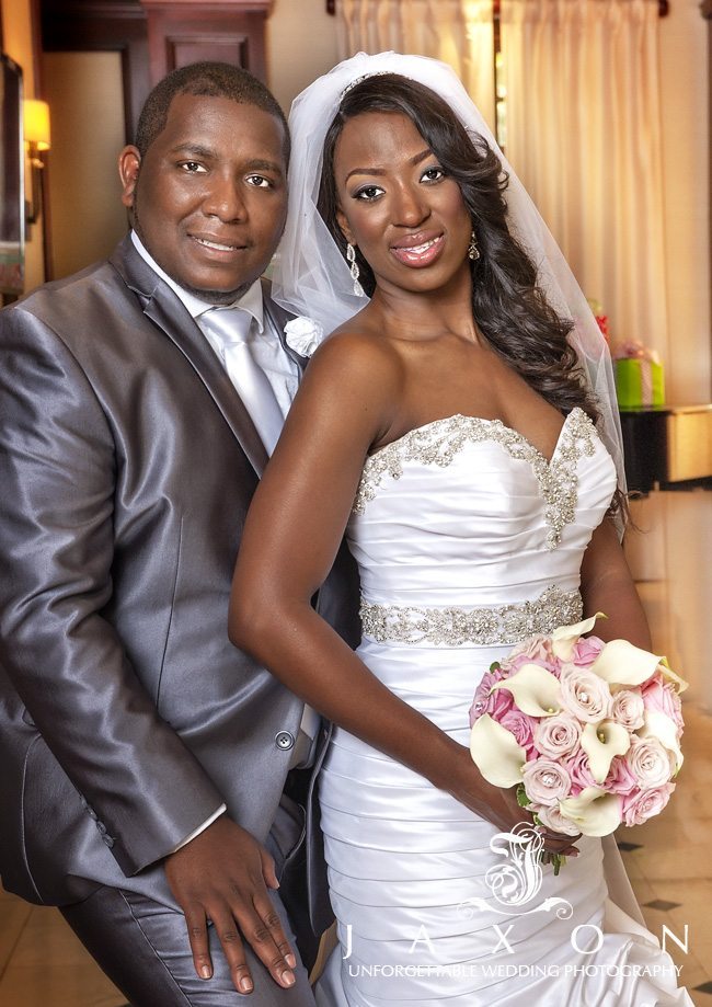 Portrait of bride and groom, he is wearing a gray suit and long silver tie, she is wearing a white trumpet dress with crystal embellished sash and neckline. Her bouquet is filled with pink and blush roses and white calla lilies at their Eagles landing wedding